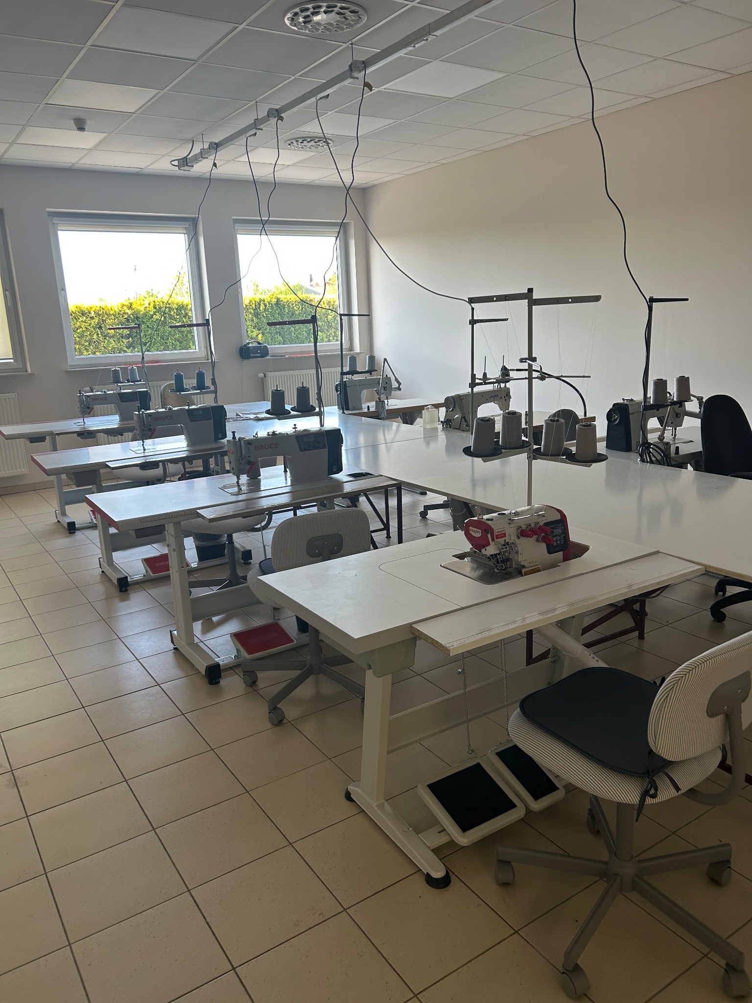 Jasne wnętrze szwalni z kilkoma stanowiskami pracy: maszyny do szycia, nici, krzesła. Widok na okna i zieleń na zewnątrz. Podłoga z płytek w odcieniach beżu.