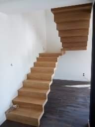 Drewniane schody dywanowe, bez balustrad, z widocznymi stopnicami i podstopnicami, prowadzące na wyższą kondygnację w jasnym wnętrzu.