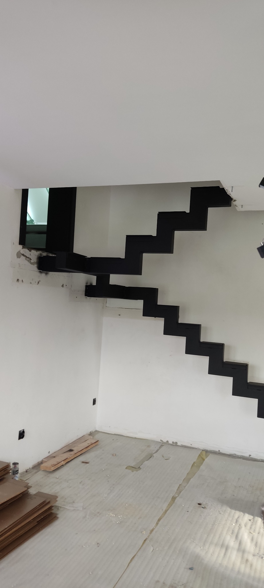 Nowoczesne, czarne schody metalowe bez balustrady, zamocowane do ściany w surowym wnętrzu. Widoczne przygotowania do wykończenia podłogi panelami.