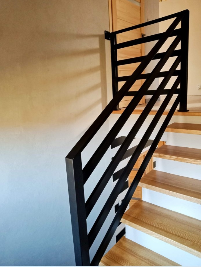 Nowoczesna balustrada schodowa z czarnych, prostokątnych profili, zamontowana na drewnianych schodach w jasnym wnętrzu. Minimalistyczny design.