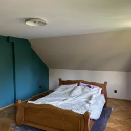 Pok&oacute;j na poddaszu po malowaniu ścian: zielona ściana kontrastuje z beżową, drewniane ł&oacute;żko z białą kołdrą, parkiet w jodełkę, okno z widokiem na zieleń.