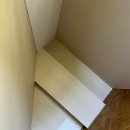 Mateusz Waligóra - Pomieszczenie po malowaniu ścian na beżowo, widoczny fragment podłogi z parkietu w jodełkę i biała klapa rewizyjna w ścianie, oświetlone naturalnym światłem.