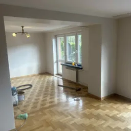 Jasne, przestronne wnętrze po remoncie z parkietem w jodełkę, widoczne narzędzia malarskie i odkurzacz. Świeżo pomalowane ściany i okno z widokiem na zieleń.