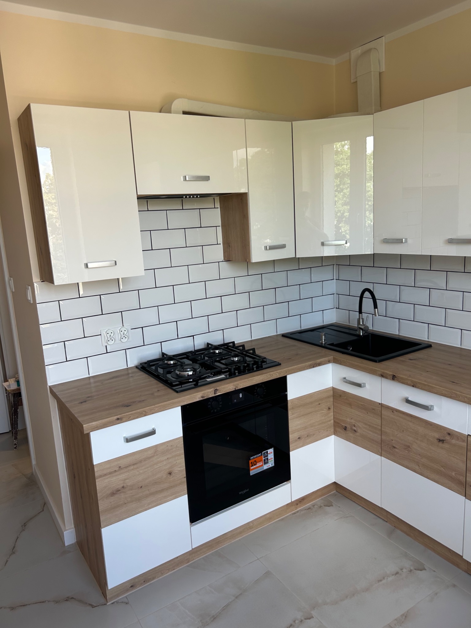 Biało-drewniana kuchnia z czarnym zlewem i płytą gazową na tle białych płytek z czarną fugą. Nowoczesny design, jasne wnętrze, widok na fragment okna.