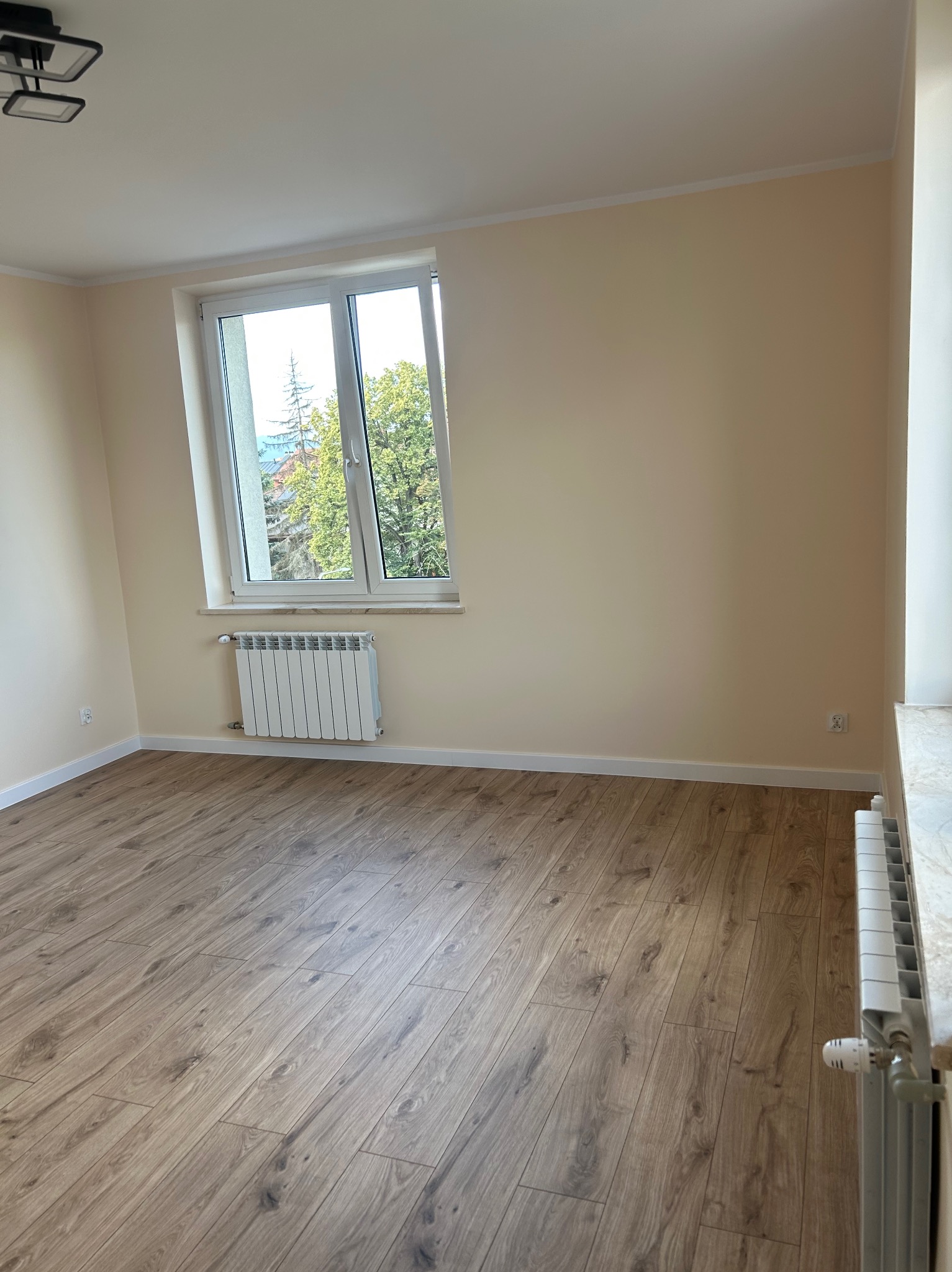 Jasne wnętrze po remoncie: drewnopodobna podłoga, kremowe ściany, białe okno z widokiem na zieleń. Grzejnik pod oknem, nowy, biały. Minimalistyczny design.