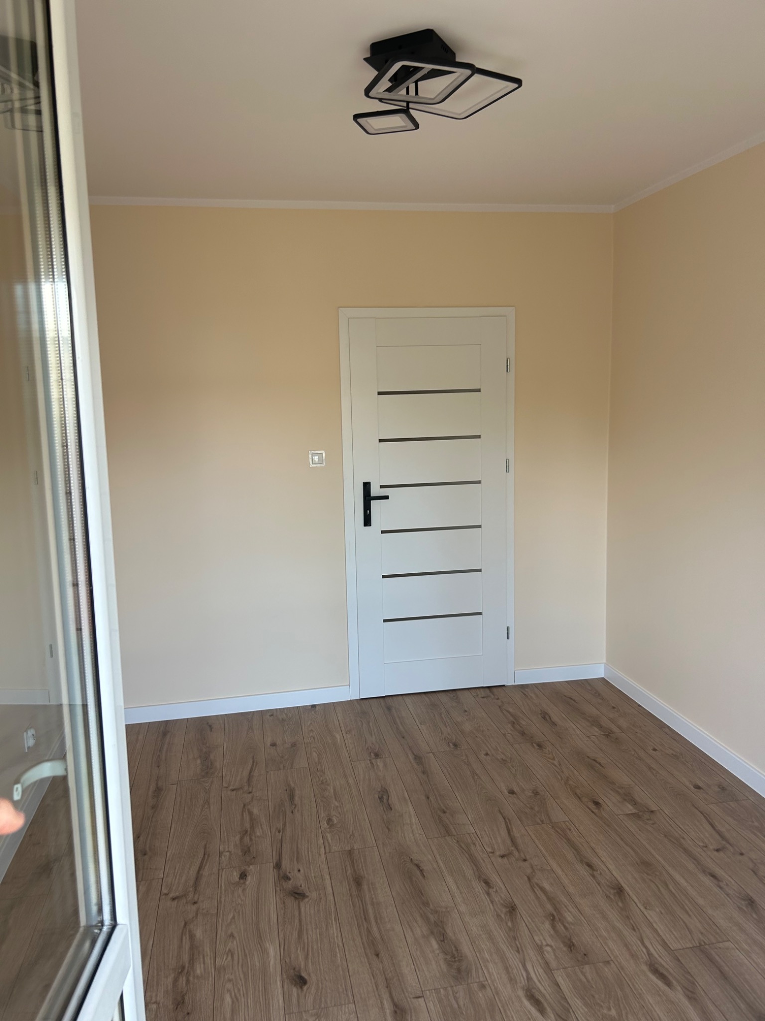 Jasne wnętrze po remoncie: drewnopodobne panele, białe listwy przypodłogowe i drzwi z poziomymi przeszkleniami. Nowoczesna lampa sufitowa.