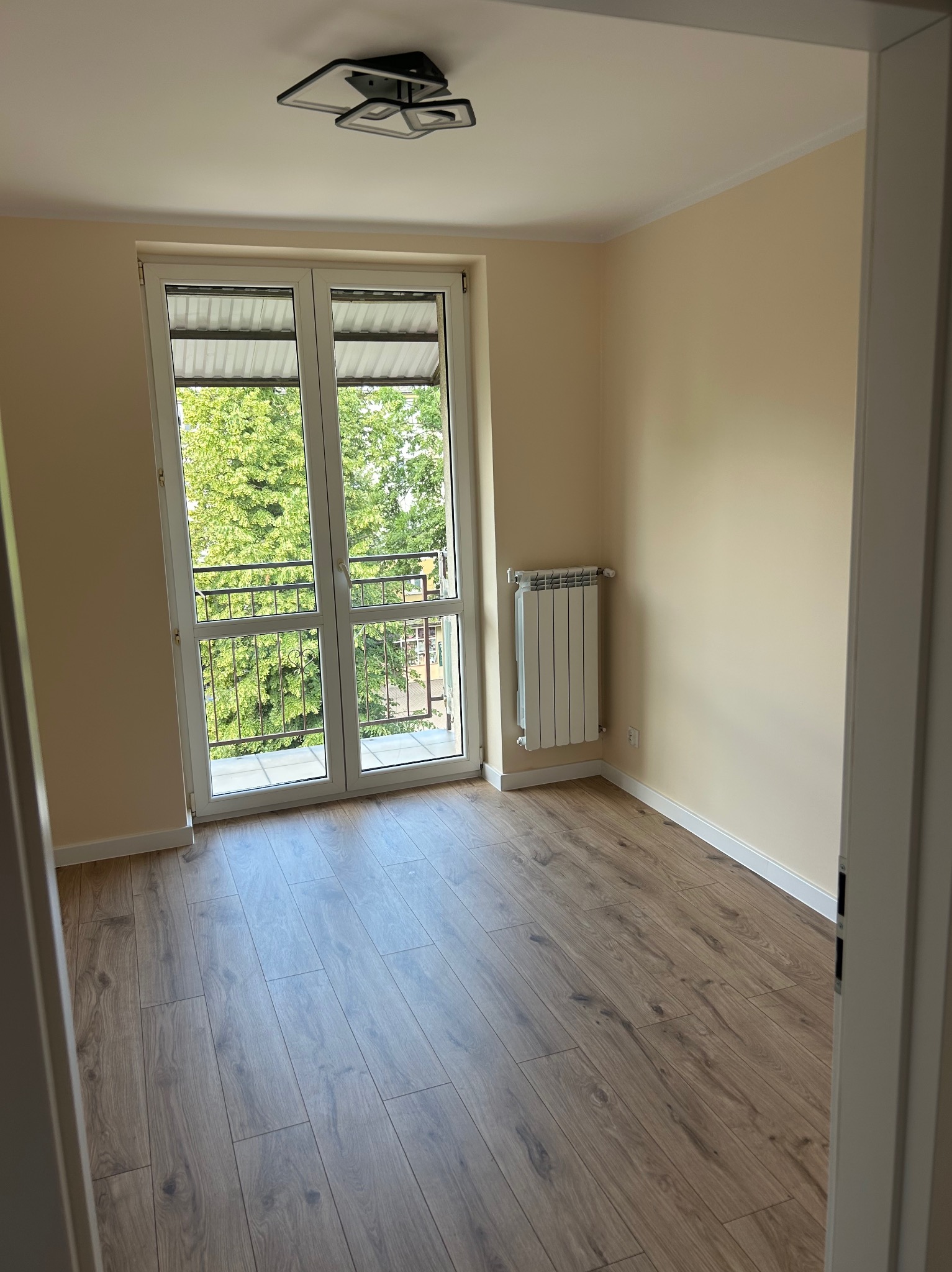 Jasne, odnowione wnętrze pokoju z drewnianą podłogą, białymi listwami przypodłogowymi i grzejnikiem. Balkon z widokiem na zieleń w tle. Nowoczesna lampa sufitowa.