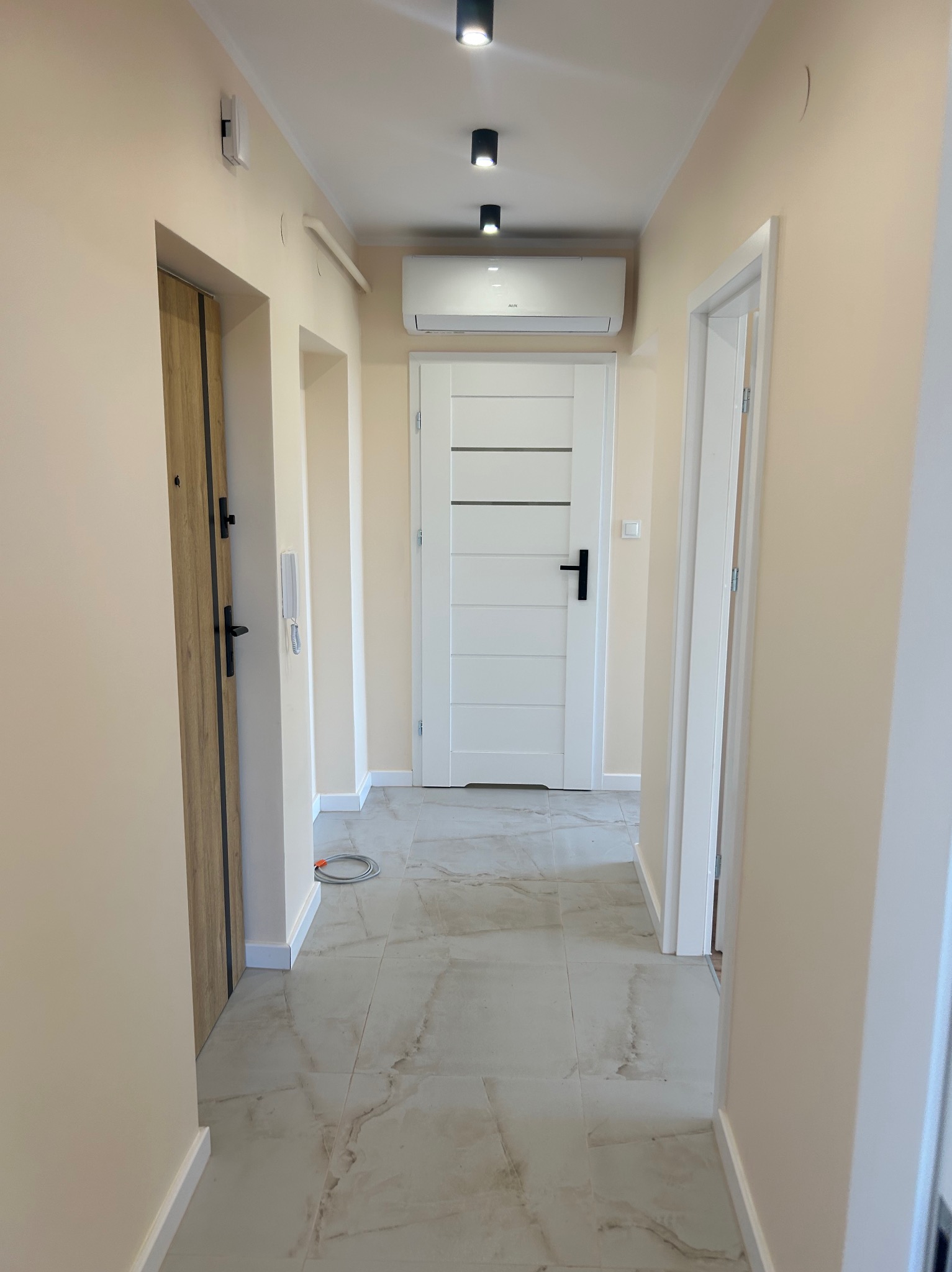 Wykończony korytarz z marmurową podłogą, białymi drzwiami z czarnymi klamkami i nowoczesnym oświetleniem punktowym. Widoczny klimatyzator nad drzwiami.