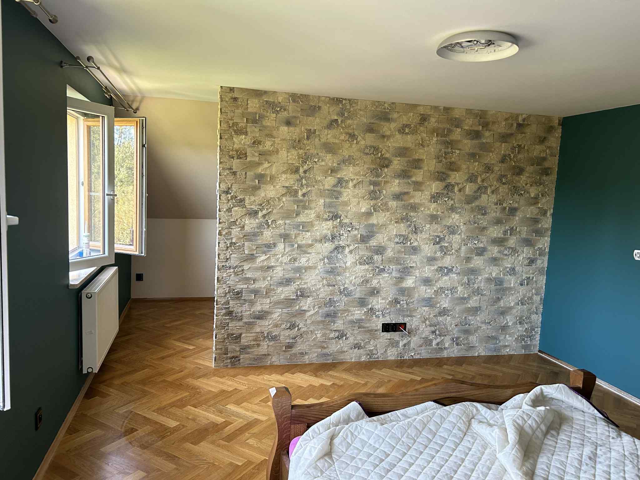 Ściana z dekoracyjnej cegły w sypialni z drewnianą podłogą w jodełkę i fragmentem łóżka. Widoczne okno i grzejnik. Ciemnozielona ściana po lewej i niebieska po prawej.