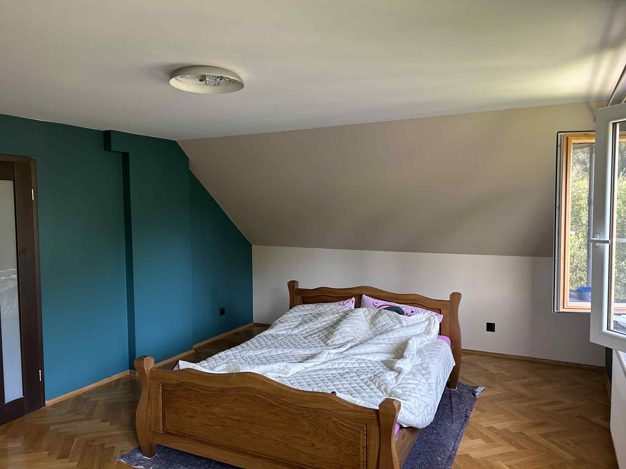 Pokój na poddaszu po malowaniu ścian: zielona ściana kontrastuje z beżową, drewniane łóżko z białą kołdrą, parkiet w jodełkę, okno z widokiem na zieleń.