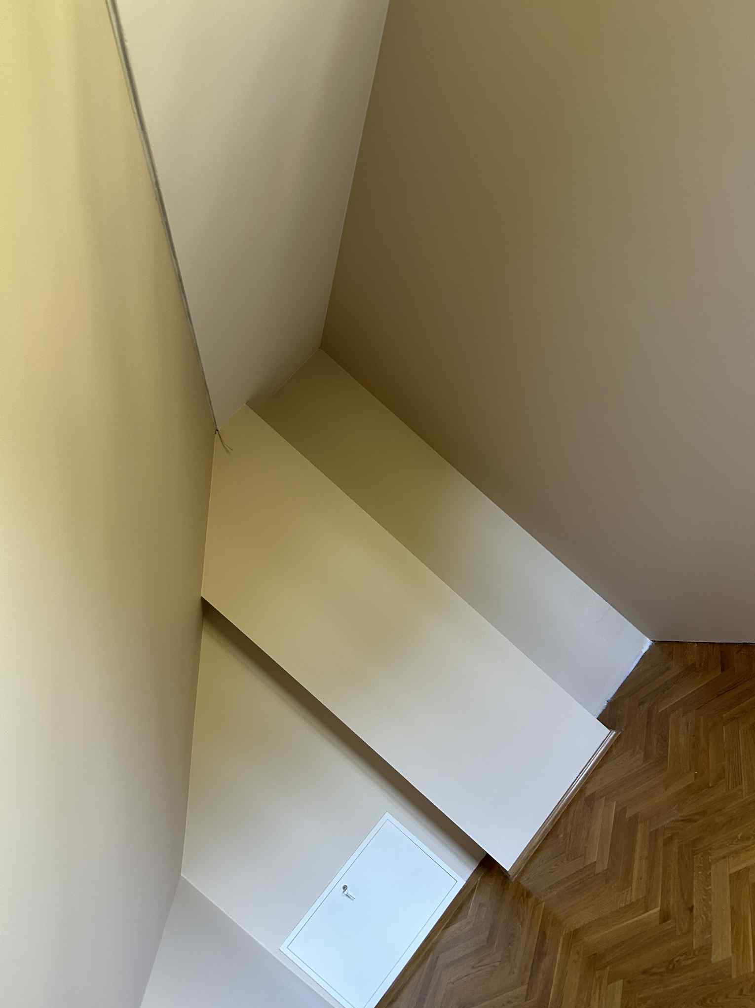 Pomieszczenie po malowaniu ścian na beżowo, widoczny fragment podłogi z parkietu w jodełkę i biała klapa rewizyjna w ścianie, oświetlone naturalnym światłem.