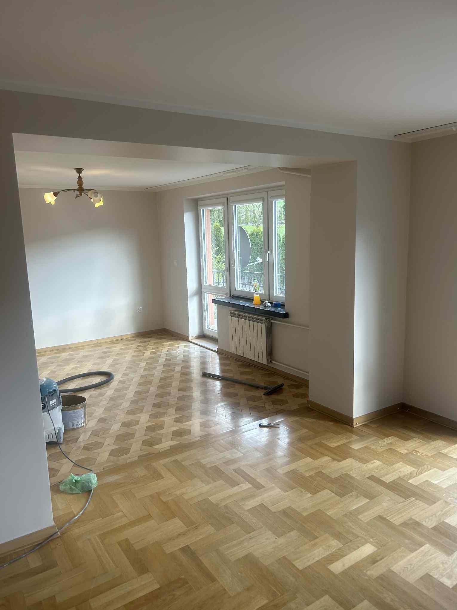 Jasne parkiety w odnowionym pokoju z białymi ścianami i oknem. Widoczne narzędzia i sprzęt po remoncie. Świeżo pomalowane ściany, gotowe do dalszego wykończenia.