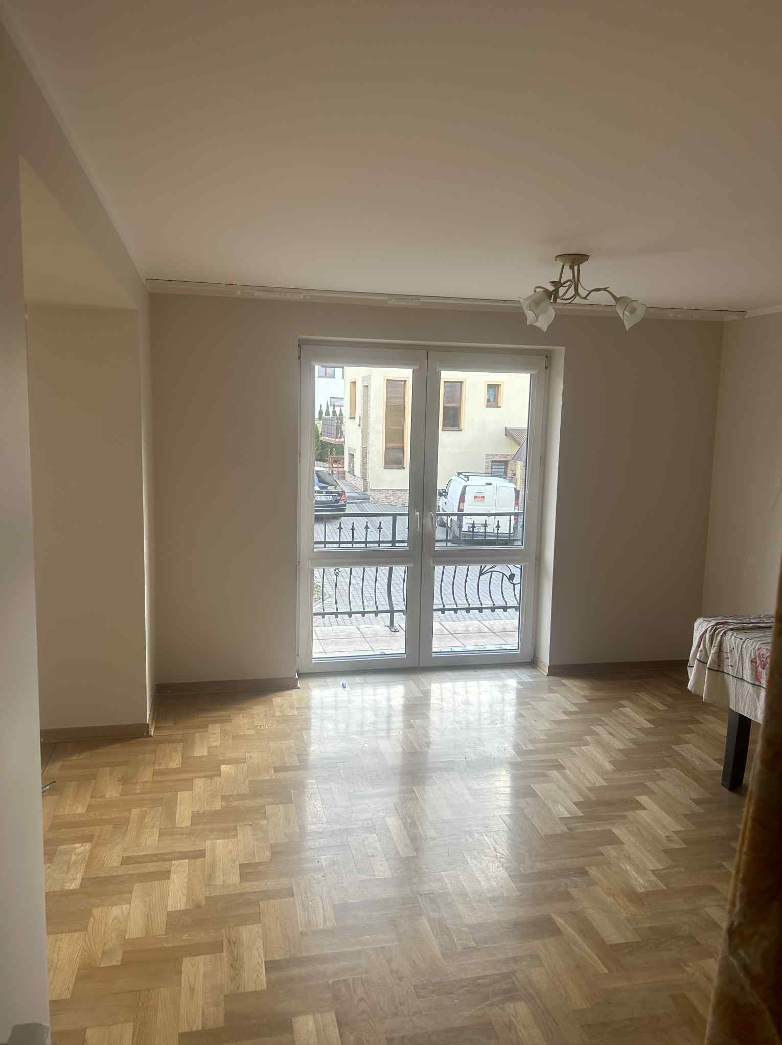 Jasne, puste wnętrze z parkietem w jodełkę, balkonem z czarną balustradą i widokiem na ulicę z zaparkowanym samochodem i białym vanem.