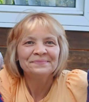 Portret uśmiechniętej kobiety w Miliczu. Blond włosy, pomarańczowa bluzka, jasna karnacja. Ujęcie od przodu, widoczne ramiona i fragment okna w tle.