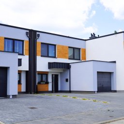 F.U.H. POL-BUD Okna Michalski - Nowoczesny dom szeregowy z grafitowymi bramami garażowymi i oknami z drewnianymi panelami. Szara kostka brukowa przed budynkiem.