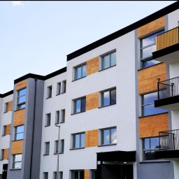 F.U.H. POL-BUD Okna Michalski - Nowoczesny budynek wielorodzinny z balkonami i oknami, wykończony elementami drewnopodobnymi, w neutralnej kolorystyce. Widok z dołu na elewację.