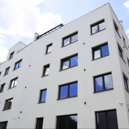 F.U.H. POL-BUD Okna Michalski - Biały budynek wielorodzinny z ciemnymi oknami, widziany z dołu, z odbiciem nieba w szybach. Minimalistyczna architektura, jasne tło nieba.