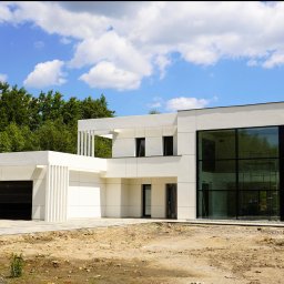 F.U.H. POL-BUD Okna Michalski - Nowoczesny, biały dom z dużymi przeszkleniami i bramą garażową. Widoczne rusztowania wewnątrz. Błękitne niebo z chmurami w tle.