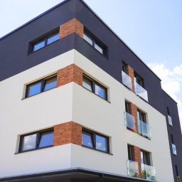 F.U.H. POL-BUD Okna Michalski - Nowoczesny budynek wielorodzinny z oknami w czarnych ramach i balkonami ze szklanymi balustradami w Tarnowskich Górach. Elewacja w kolorach bieli i grafitu.