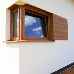 F.U.H. POL-BUD Okna Michalski - Brązowe okno z widokiem na niebo i chmury, zamontowane na białej ścianie budynku z elementami dekoracyjnymi w kolorze drewna, widok z zewnątrz.