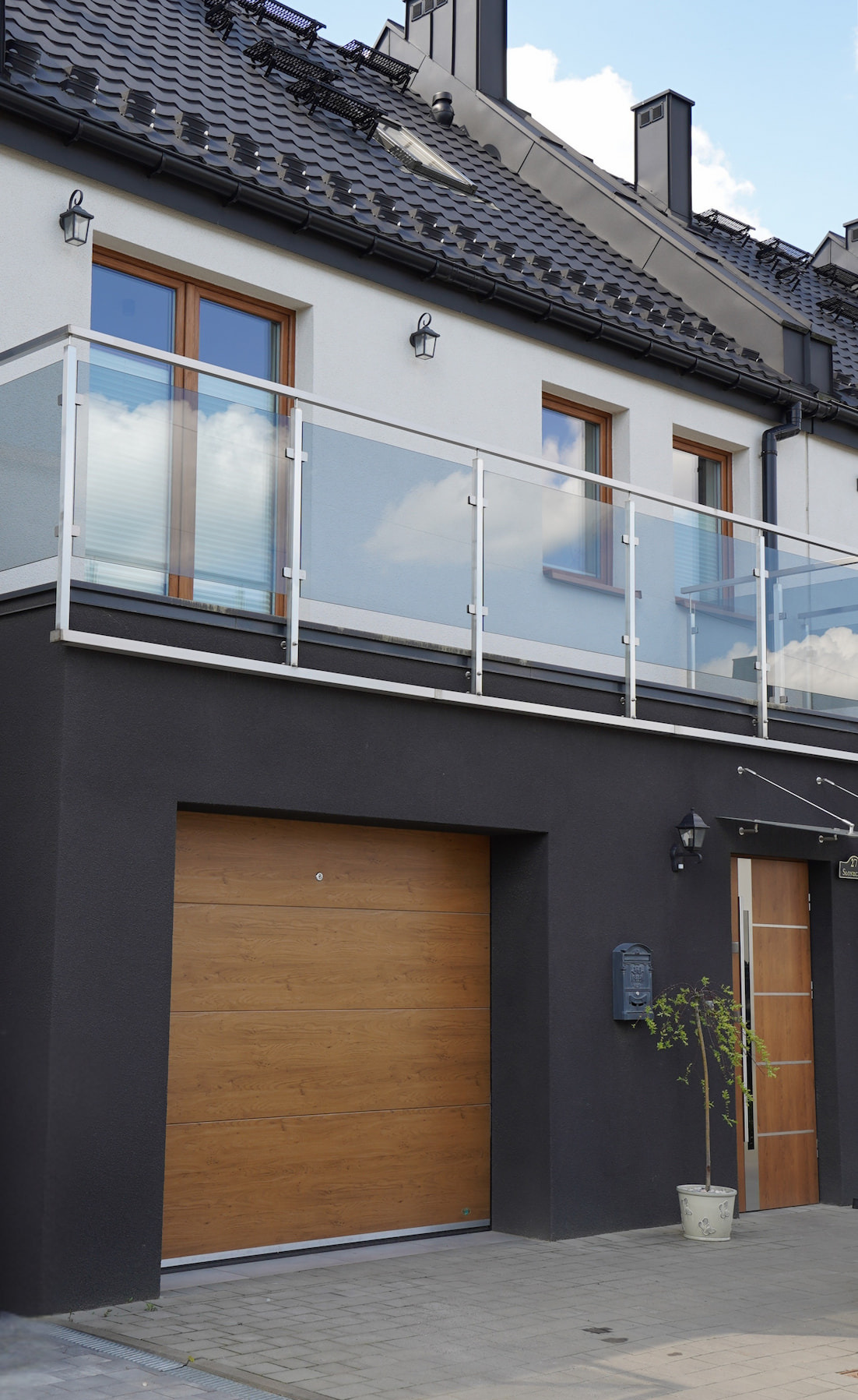 Nowoczesny dom z drewnianą bramą garażową i szklaną balustradą balkonu. Elewacja w bieli i graficie, czarny dach. Drzwi wejściowe z boku, roślina w donicy.