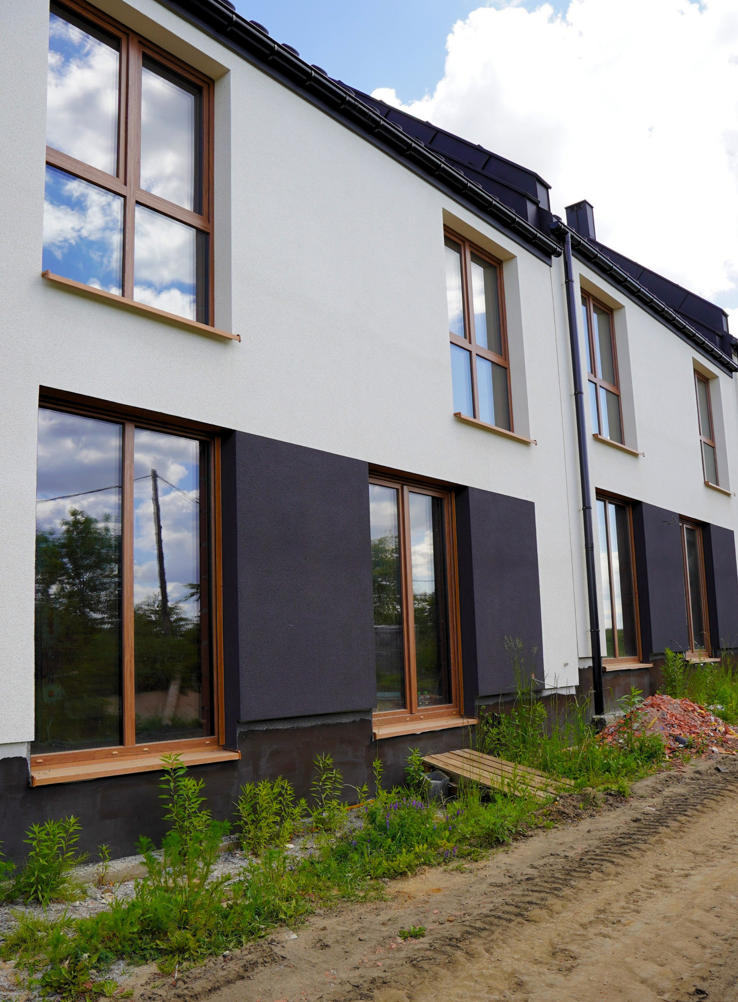 Elewacja domu z drewnianymi oknami, widoczne odbicie nieba w szybach. Minimalistyczny design, biała ściana i ciemnoszara podmurówka. Świeża zieleń przed budynkiem.