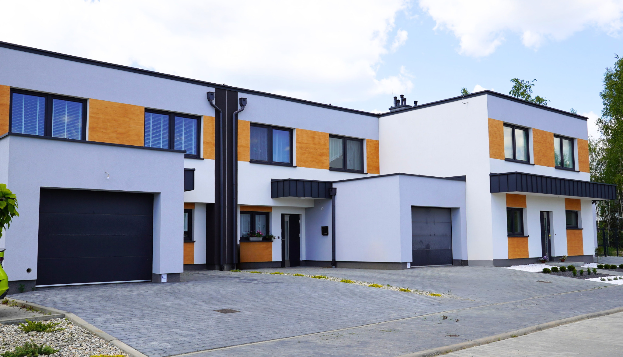 Nowoczesny dom szeregowy z grafitowymi bramami garażowymi i oknami z drewnianymi panelami. Szara kostka brukowa przed budynkiem.