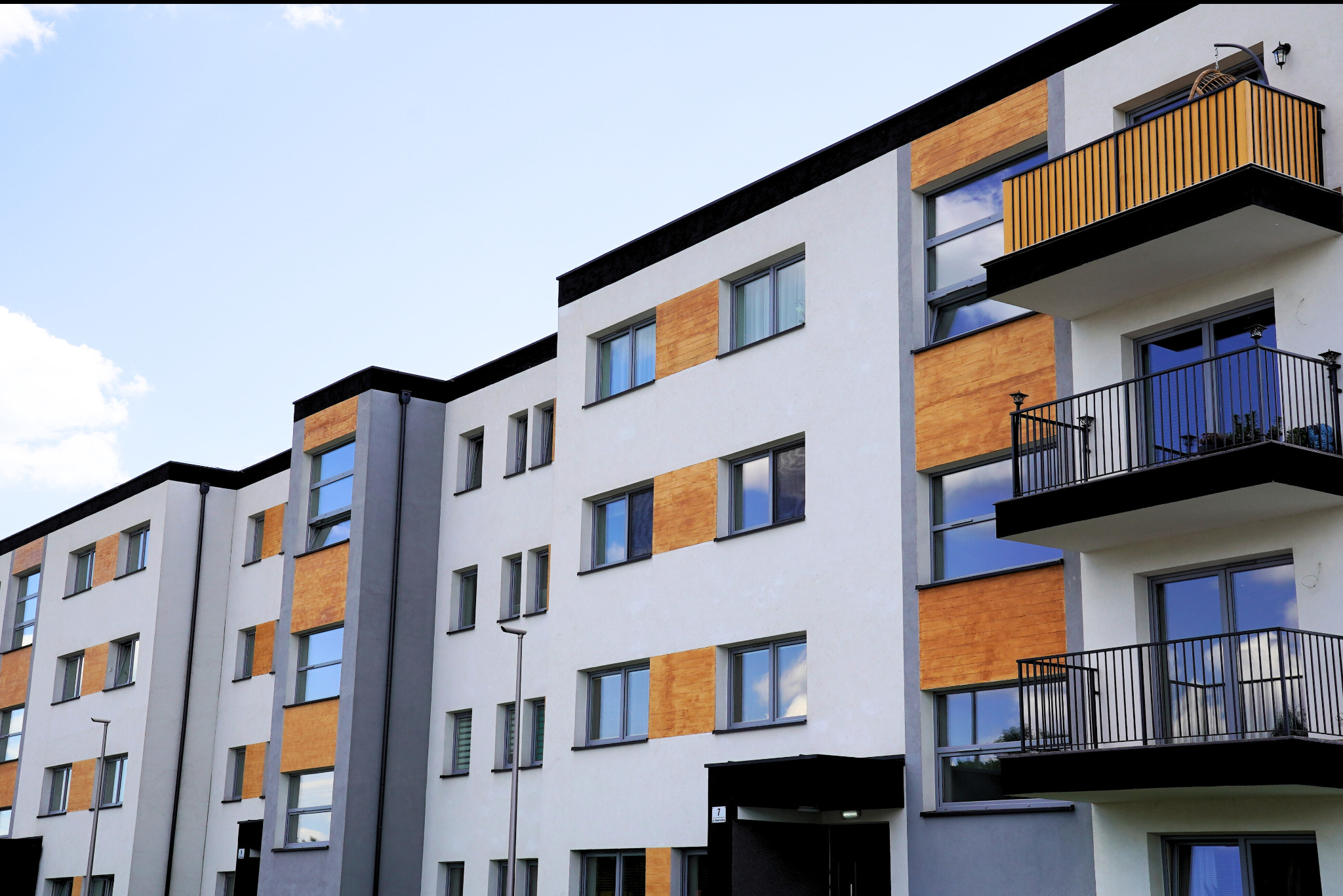 Nowoczesny budynek wielorodzinny z balkonami i oknami, wykończony elementami drewnopodobnymi, w neutralnej kolorystyce. Widok z dołu na elewację.