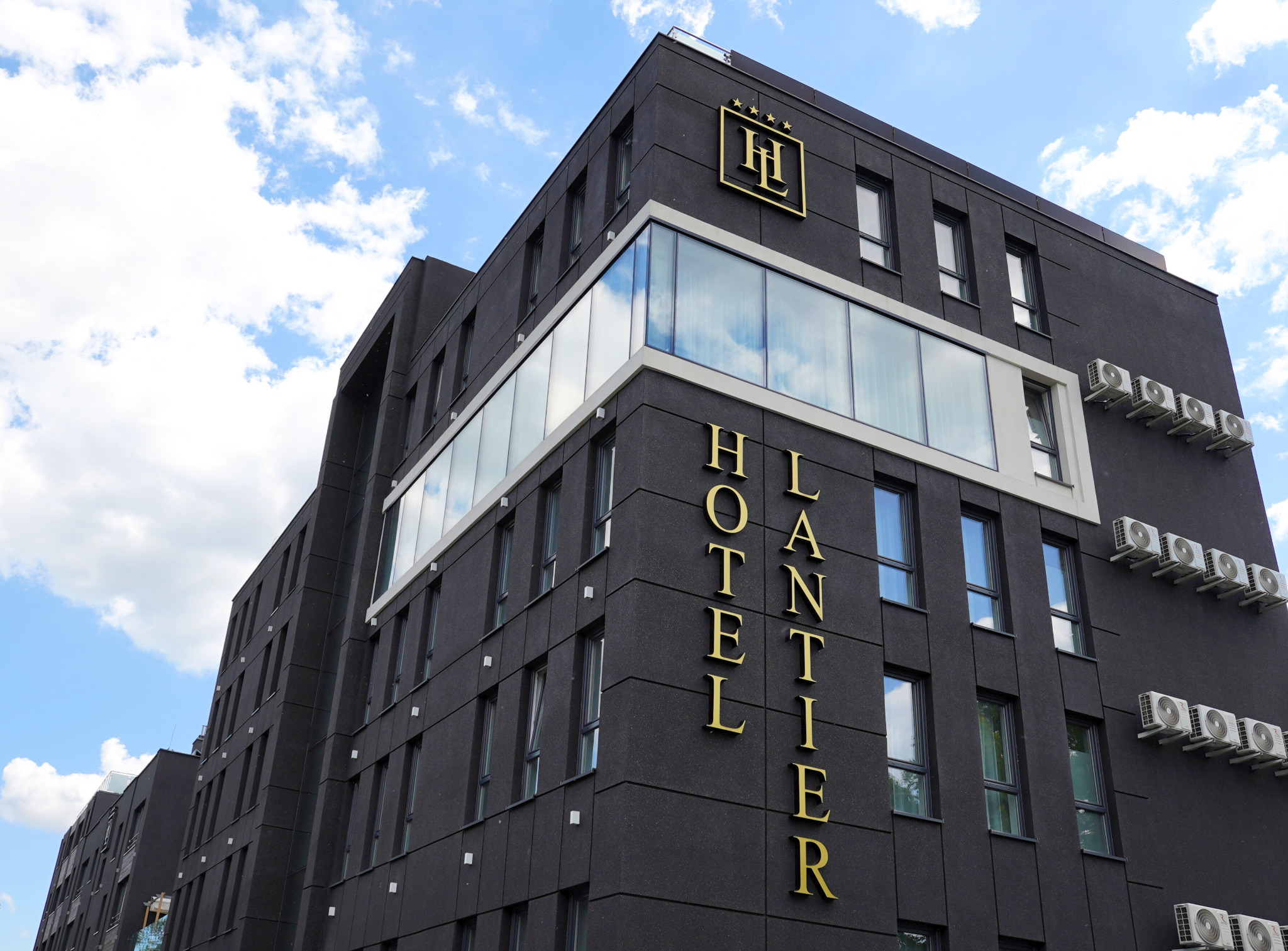 Nowoczesny budynek hotelu z ciemną elewacją, dużymi oknami i złotym napisem HOTEL LANTIER, widziany z dołu na tle błękitnego nieba z chmurami.