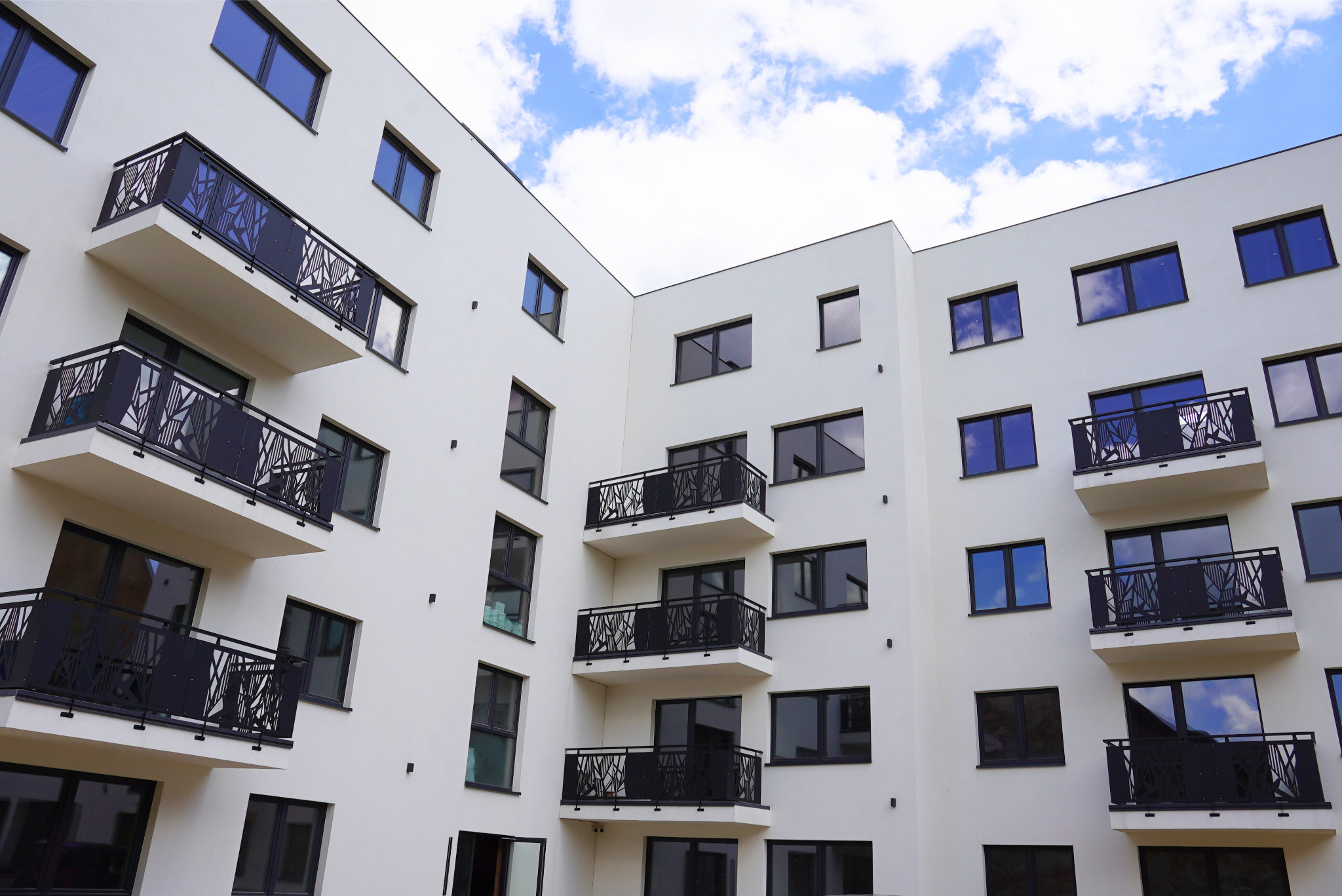 Biały budynek wielorodzinny z balkonami o czarnych, ażurowych balustradach. Widok z dołu na fasadę z oknami i fragmentem błękitnego nieba z chmurami.