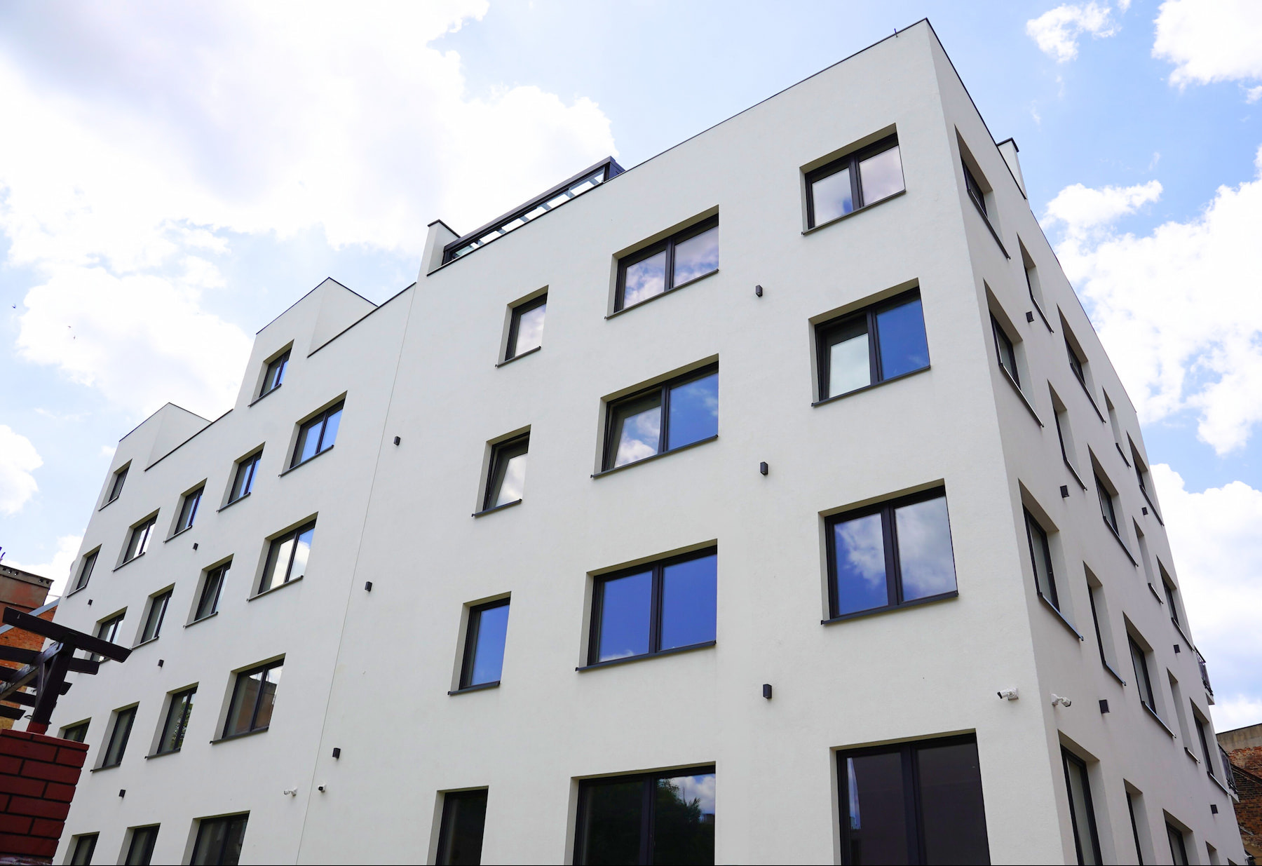 Biały budynek wielorodzinny z ciemnymi oknami, widziany z dołu, z odbiciem nieba w szybach. Minimalistyczna architektura, jasne tło nieba.