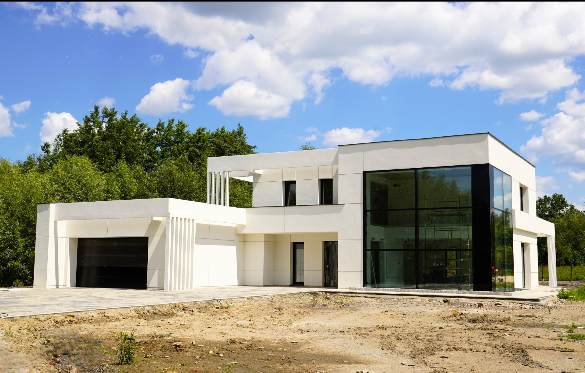 Nowoczesny, biały dom z dużymi przeszkleniami i bramą garażową. Widoczne rusztowania wewnątrz. Błękitne niebo z chmurami w tle.