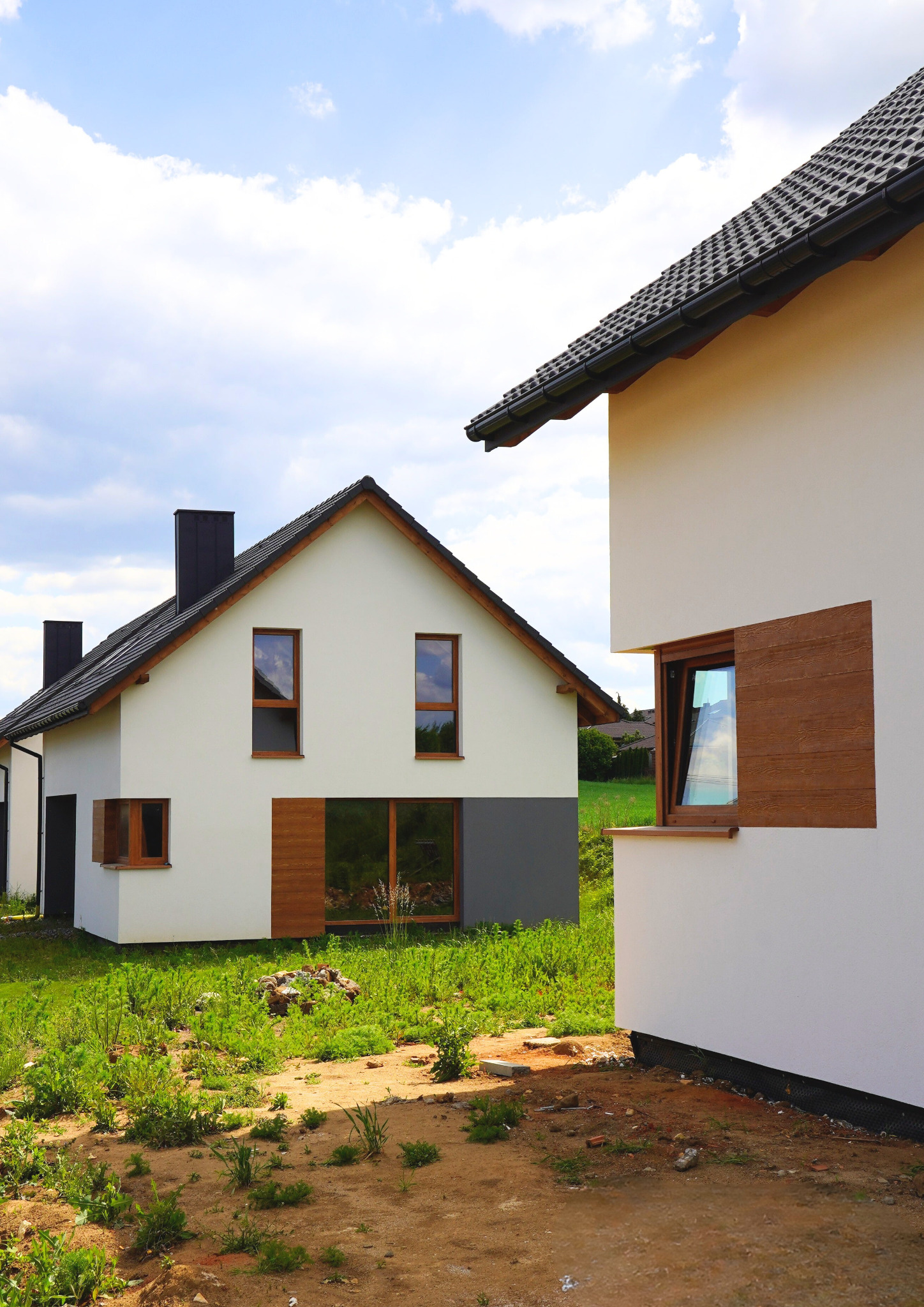 Dwa nowoczesne domy jednorodzinne z drewnianymi ramami okiennymi, białą elewacją i ciemnymi dachami, otoczone zielenią. Jeden dom z szarą podstawą.
