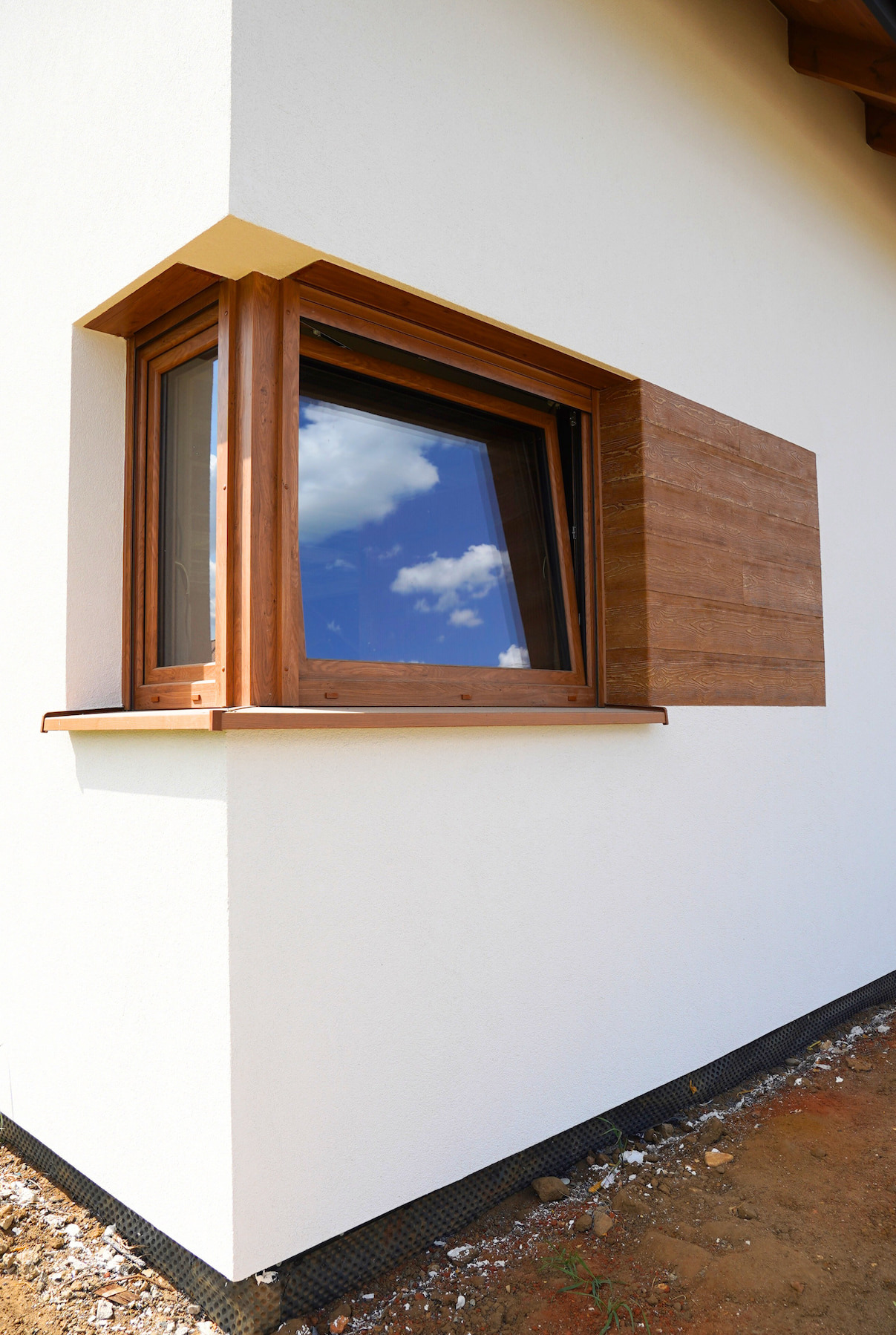 Brązowe okno z widokiem na niebo i chmury, zamontowane na białej ścianie budynku z elementami dekoracyjnymi w kolorze drewna, widok z zewnątrz.