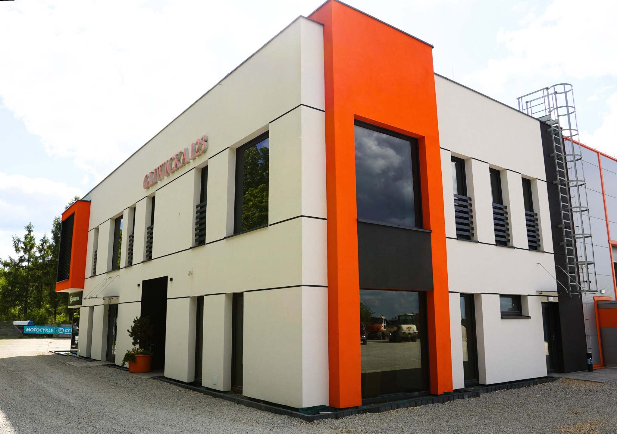 Nowoczesny budynek z oknami i pomarańczowymi akcentami, widoczny z zewnątrz. Minimalistyczna architektura, duże przeszklenia, elewacja w jasnych kolorach.