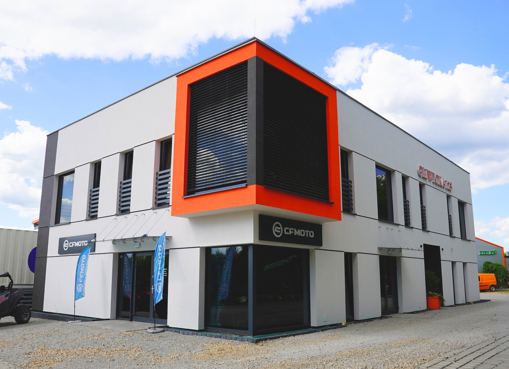 Nowoczesny budynek biurowy z dużymi oknami i pomarańczowymi akcentami architektonicznymi, widoczny z zewnątrz w słoneczny dzień.