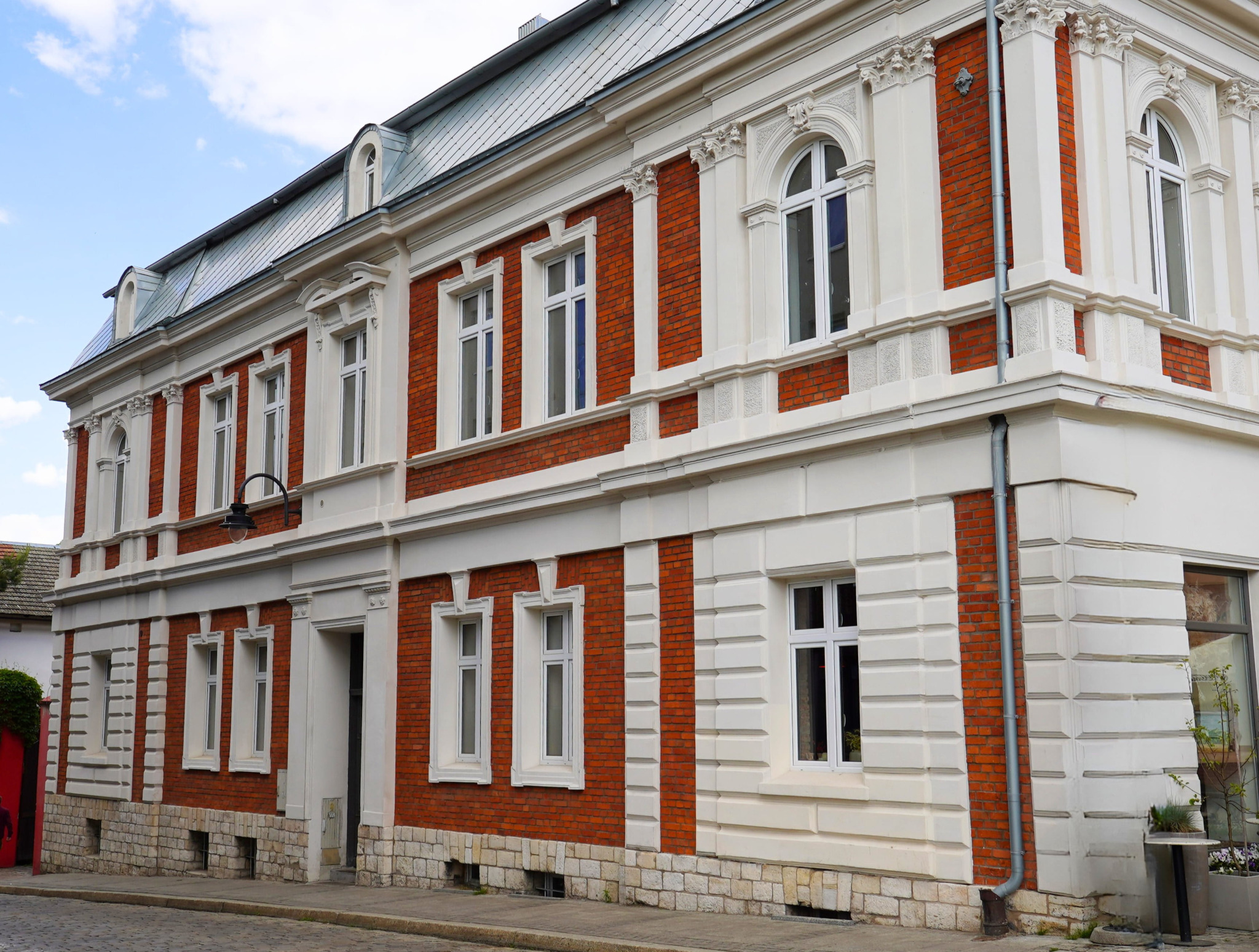 Elewacja dwukondygnacyjnego budynku z czerwonej cegły i białymi elementami dekoracyjnymi. Widoczne okna z białymi ramami i metalowy dach.