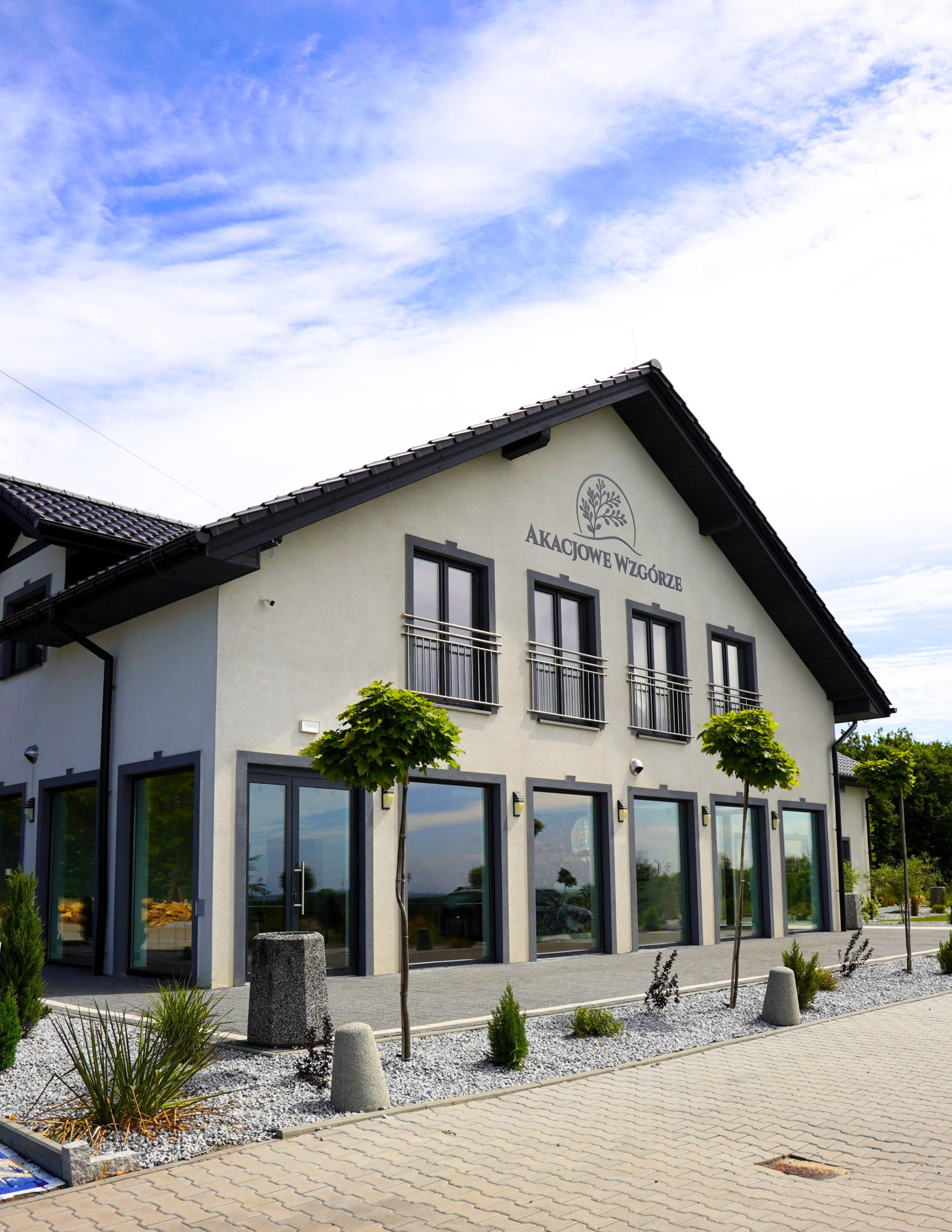 Elewacja budynku 'Akacjowe Wzgórze' z ciemnymi oknami i drzwiami. Przed budynkiem małe drzewka i kamienne rabaty.