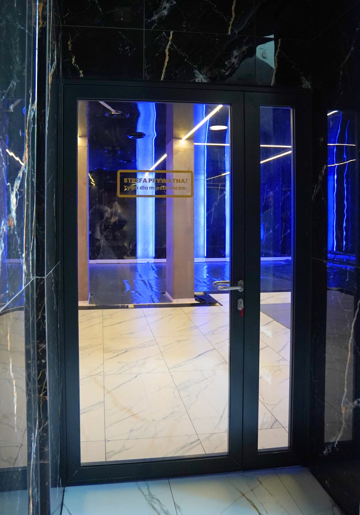 Szklane drzwi z czarną ramą, przez które widać nowoczesne wnętrze z niebieskim oświetleniem i marmurowymi elementami. Na drzwiach naklejka 'Strefa prywatna!'.