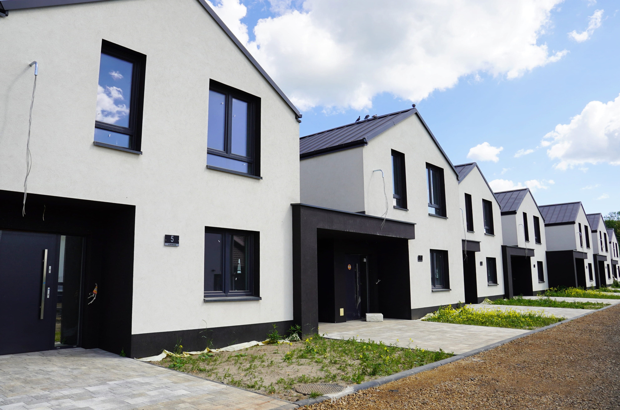 Szereg nowych domów jednorodzinnych z ciemnymi oknami i drzwiami, w jasnej kolorystyce elewacji, z widocznym numerem domu '5'.