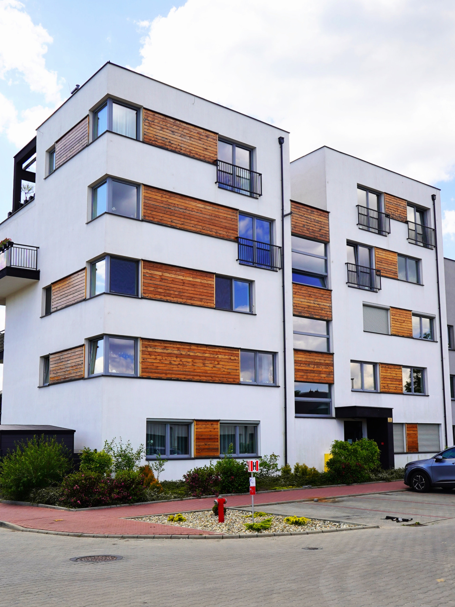 Nowoczesny budynek wielorodzinny z białymi elewacjami i drewnianymi panelami dekoracyjnymi wokół okien, balkony z czarnymi barierkami, widok z ulicy.