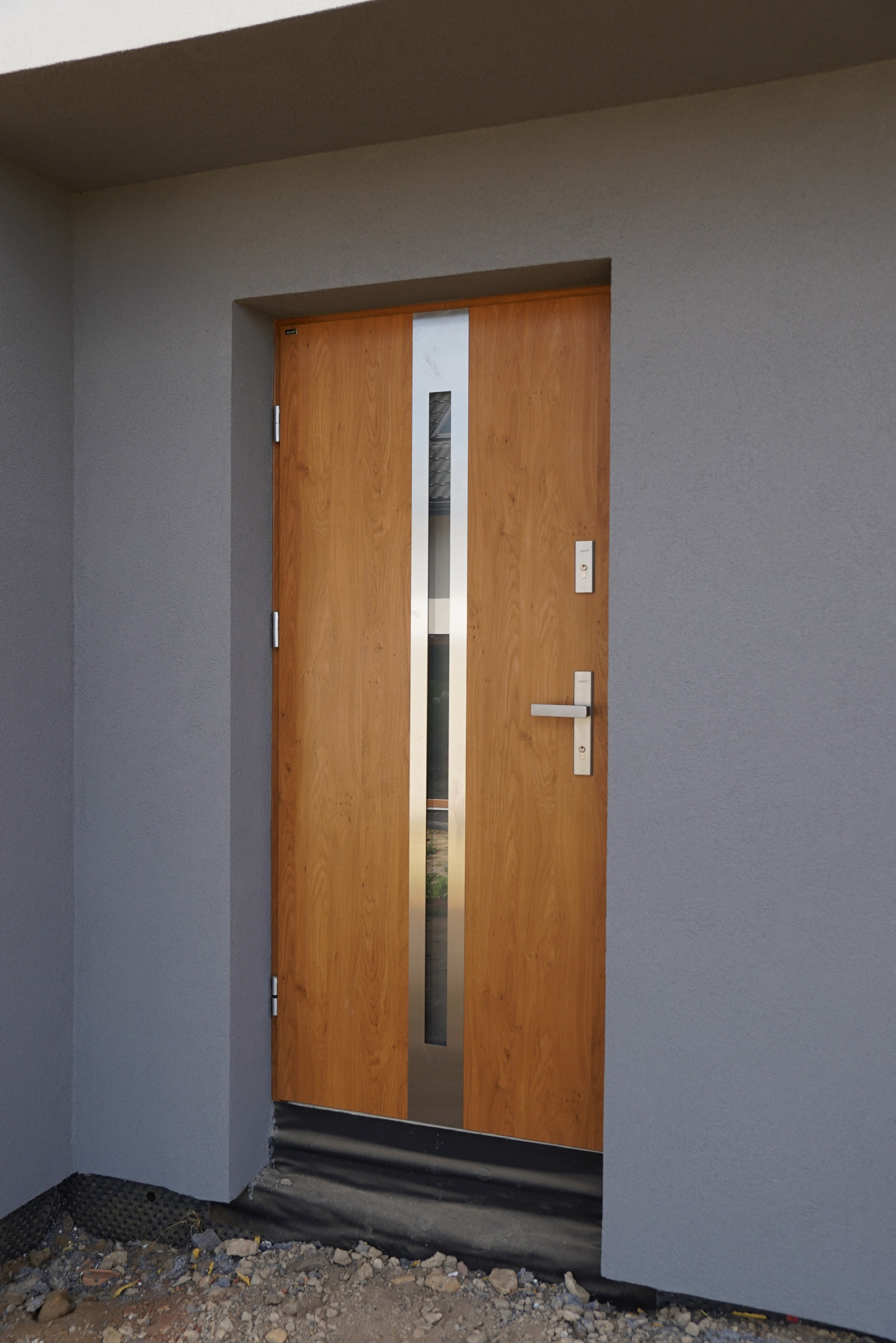 Drewnopodobne drzwi wejściowe z przeszkleniem i srebrnymi akcentami, zamontowane w szarej elewacji budynku w trakcie wykończenia. Widoczne detale montażowe.