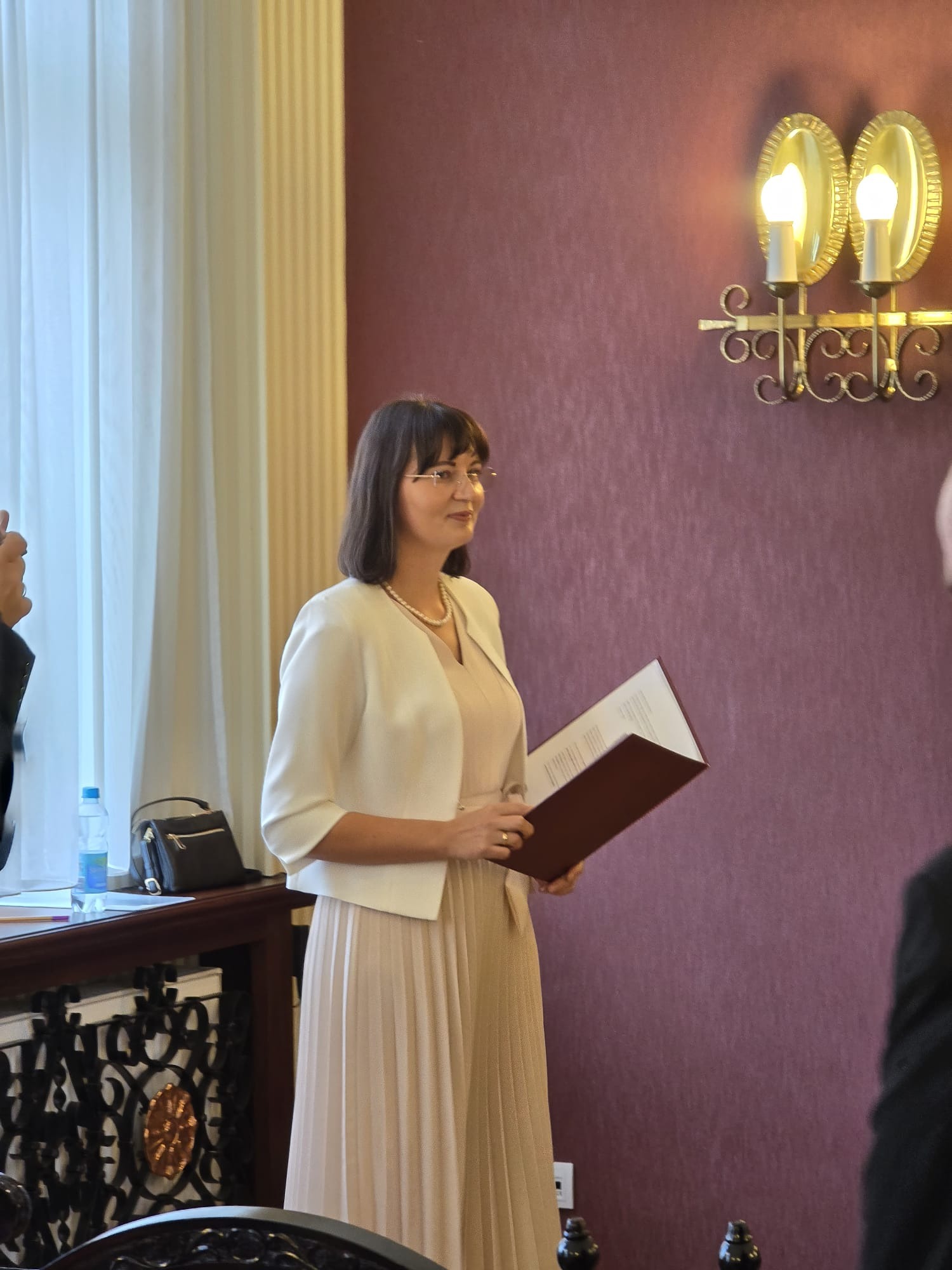 Kobieta w jasnym stroju trzyma dokument w brązowej okładce, stojąc w eleganckim wnętrzu z ozdobnym oświetleniem ściennym i zasłonami.