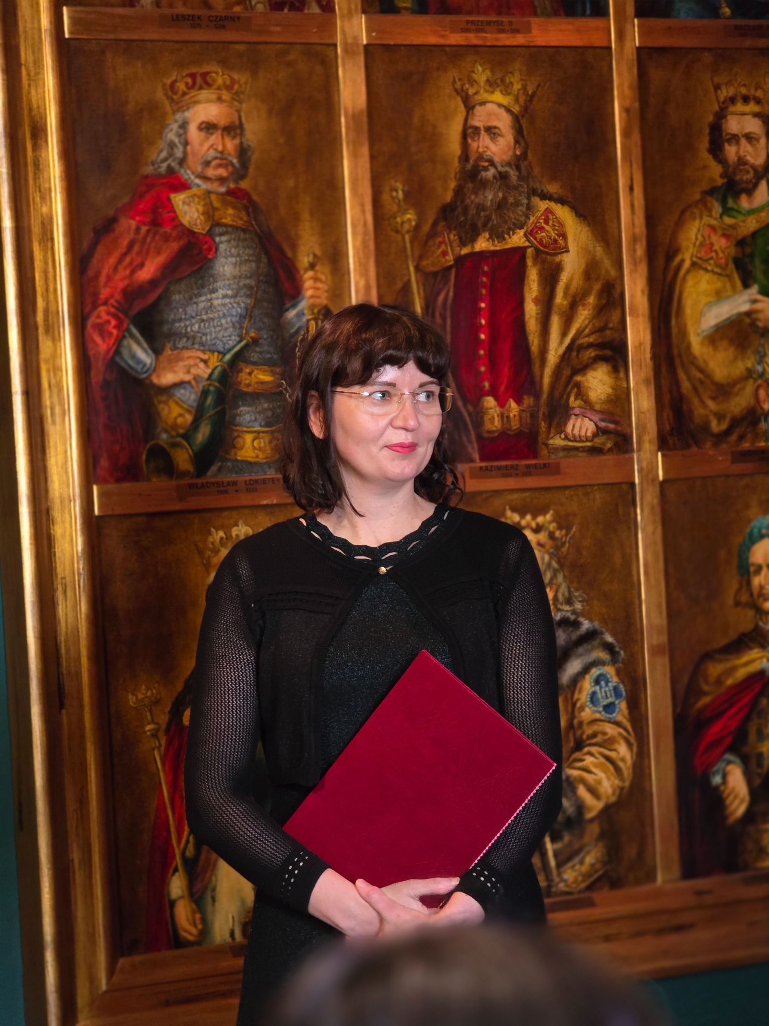 Kobieta w okularach trzyma czerwoną teczkę na tle portretów królów Polski w złotych ramach. Uśmiechnięta, patrzy w prawo.
