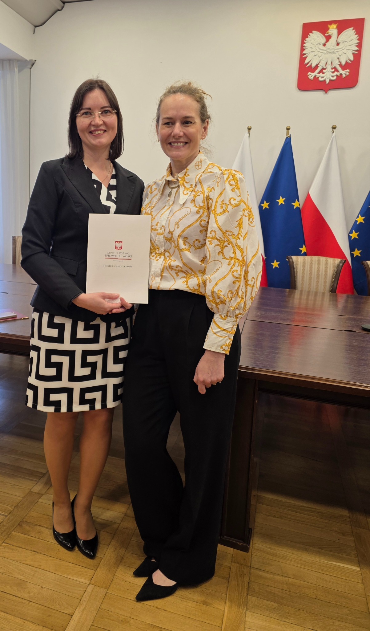 Dwie uśmiechnięte kobiety prezentują dokument Ministerstwa Sprawiedliwości na tle flag Polski i UE. Jedna w czarno-białej sukience, druga w bluzce w złote wzory.