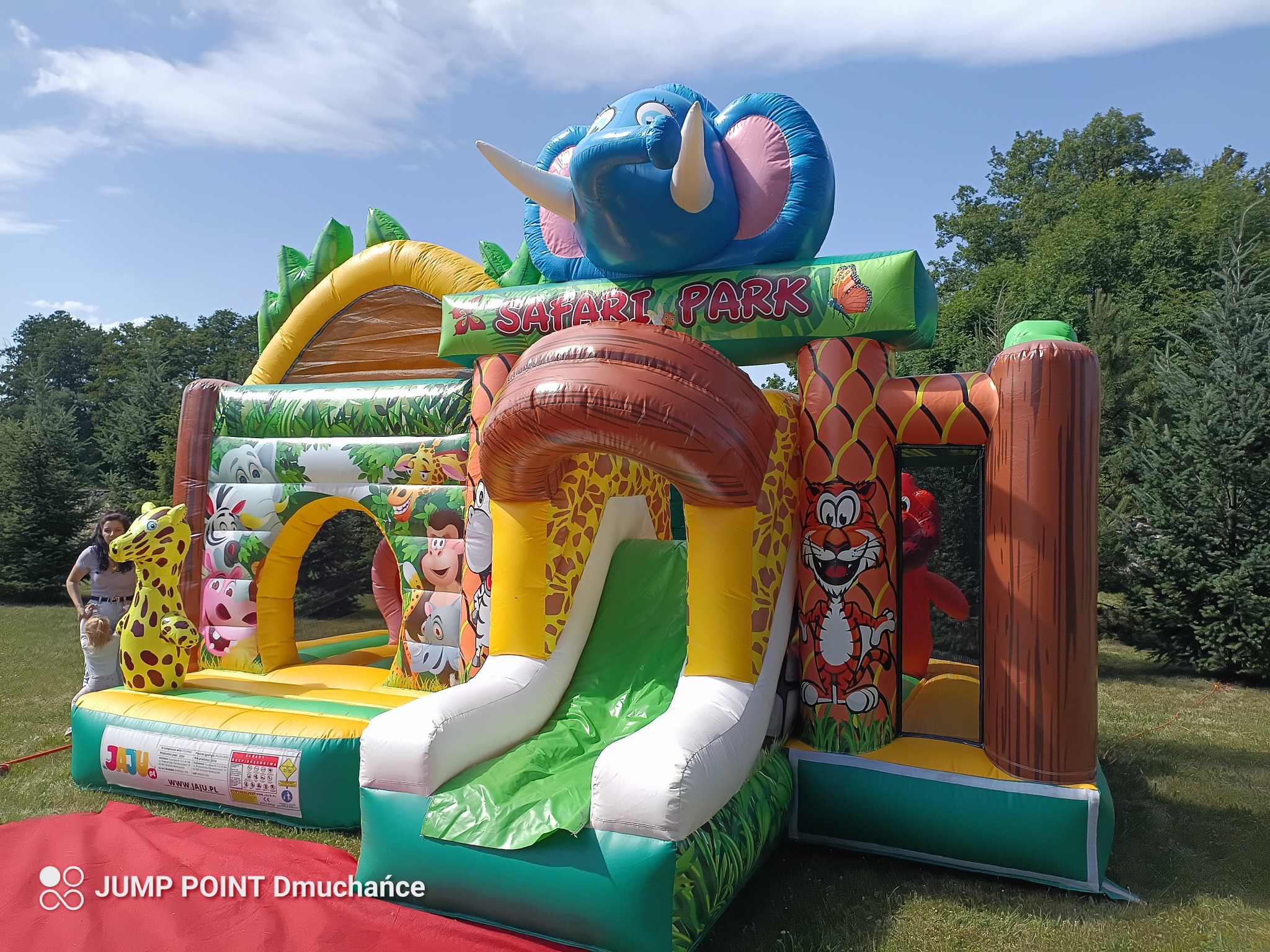 Kolorowy dmuchany park rozrywki Safari Park z motywami zwierzęcymi, zjeżdżalnią i postaciami zwierząt, ustawiony na trawie w plenerze, w tle drzewa.