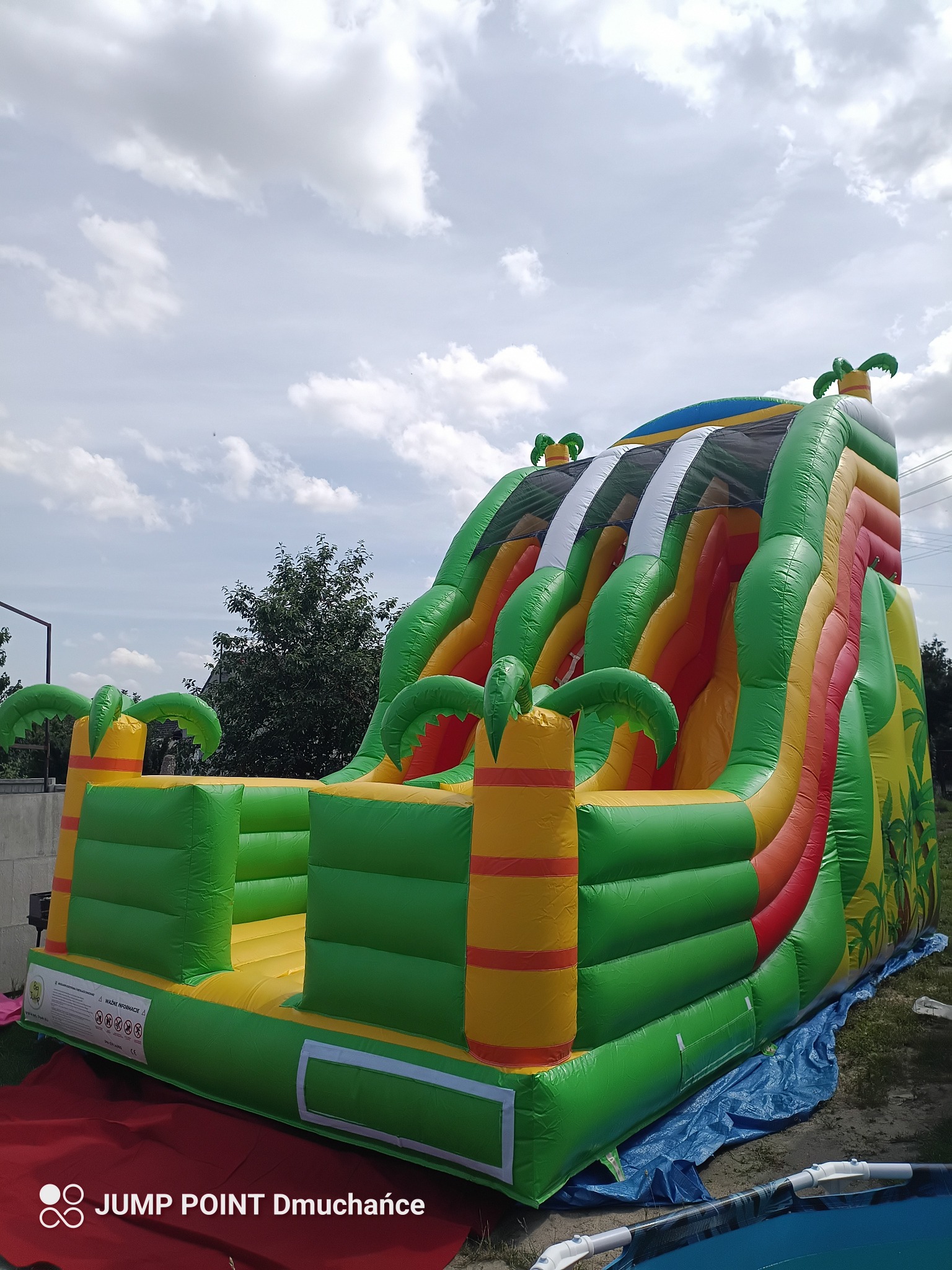 Kolorowa, dmuchana zjeżdżalnia z motywem tropikalnym i palmami, na trawie pod pochmurnym niebem. Widok z dołu, ukazujący konstrukcję i detale atrakcji.