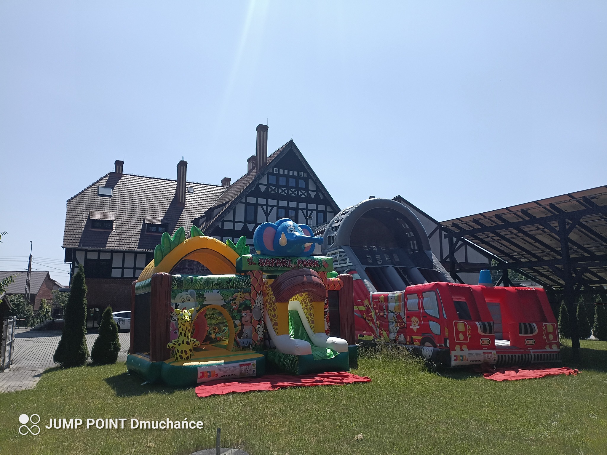 Kolorowe dmuchańce: safari z słoniem i wóz strażacki na trawie przed budynkiem z muru pruskiego w słoneczny dzień. Atrakcje dla dzieci na imprezę plenerową.
