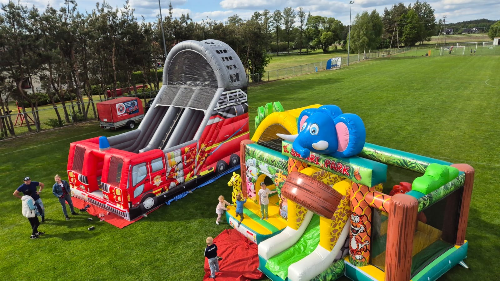 Dwa dmuchańce: straż pożarna ze zjeżdżalnią i safari park z motywem słonia. Dzieci bawią się na trawiastym terenie, widoczny także trailer z logo firmy.