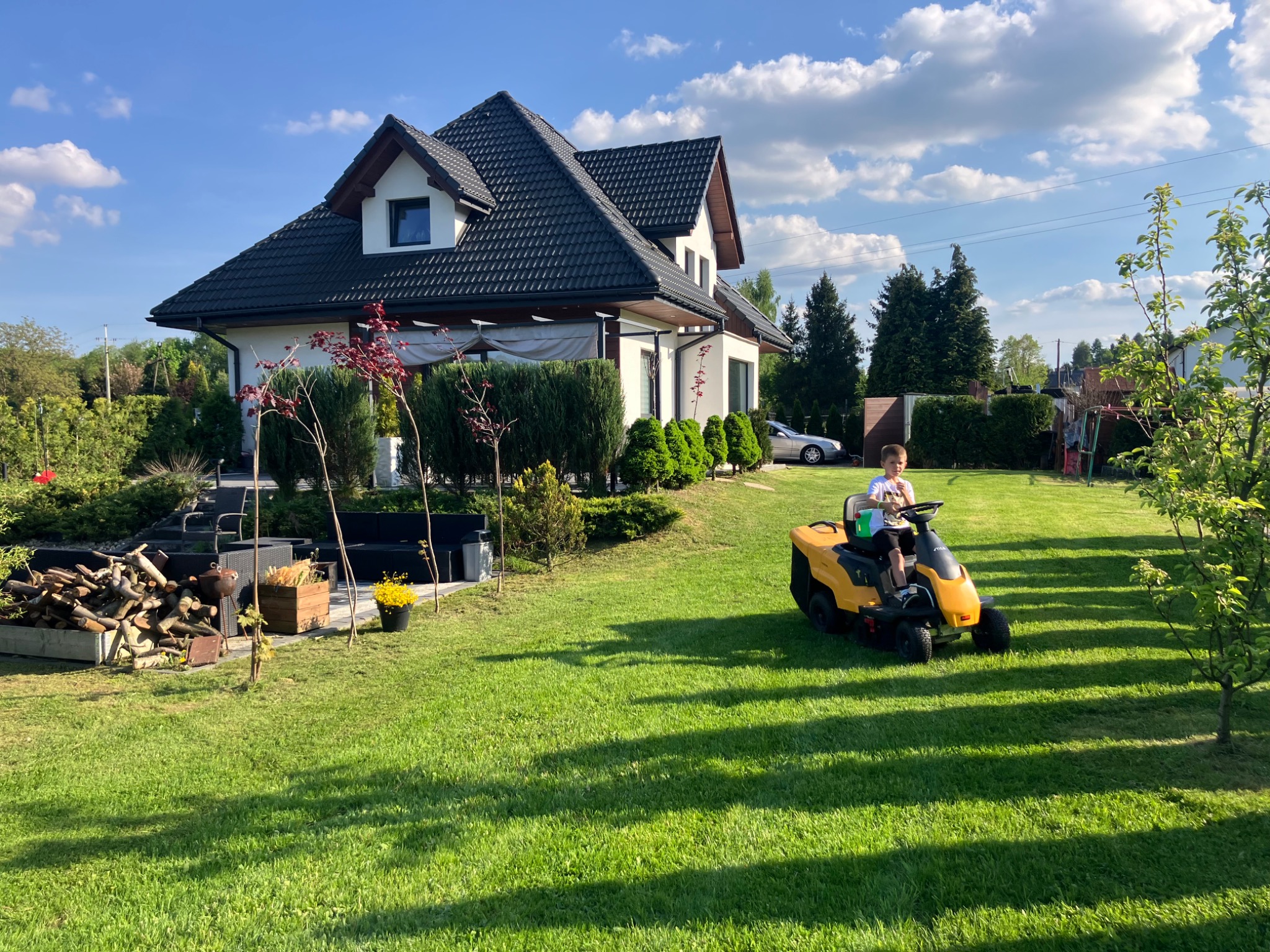 Chłopiec kosi trawę na traktorze w słonecznym ogrodzie. W tle dom z ciemnym dachem i zadbane krzewy. Widoczne ścięte drewno i dekoracje ogrodowe.